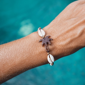Peut inclure: Un bracelet en coquille de noix de coco marron avec un charme en forme de palmier, des coquillages blancs et des perles dorées.