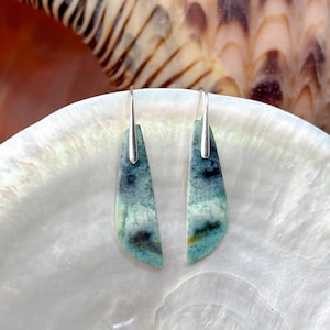 Könnte beinhalten: Ein Paar Ohrringe mit silbernen Haken und gebogenen, rechteckigen Steinen. Die Steine zeigen Blau-, Grün- und Weißtöne mit dunklen und gelben Details. Die Ohrringe werden auf einer weißen, perlmuttartigen Oberfläche präsentiert.