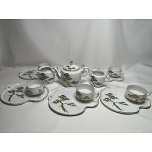 May include: A white porcelain tea set featuring a green and brown pine tree motif with silver trim. The set includes a teapot, a creamer, a sugar bowl, and several cups paired with matching plates that have a distinct curved, asymmetrical shape.