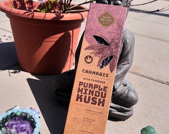 May include: A brown box of incense sticks with the text "Cannabis with Terpenes Purple Hindu Kush Indica" and "Fruit Base Incense Sticks Scented with a Blend of Fruit" on the front. The box is sitting on a table with a small statue of Buddha and a few green and blue stones.