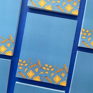 May include: A set of blue notepads with a border of yellow pasta shapes. The pasta shapes include bowtie pasta, penne pasta, tortellini, and other shapes.