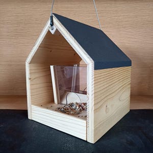 May include: A wooden bird feeder shaped like a house with a black roof. The feeder has a clear plastic tray for holding birdseed and a small opening for birds to access the food.