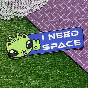 Puede incluir: Una pegatina rectangular con fondo azul y texto blanco que dice I NEED SPACE. En el lado izquierdo, hay una ilustración de dibujos animados de un personaje alienígena de color verde claro con grandes ojos negros y antenas.