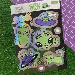May include: A sticker sheet featuring holographic stickers with a "Glorp Kitty" theme. The stickers depict green alien cat hybrids in various poses, including one in a UFO and another on a planet. The sheet also includes the text "GNARP".