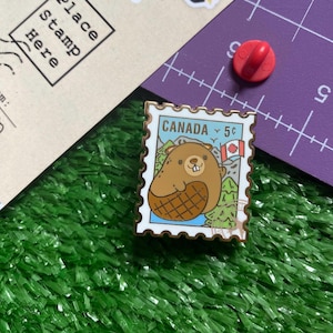 May include: A rectangular enamel pin resembling a Canadian postage stamp, depicting a cartoon beaver. The pin has a white border with the word "CANADA" and a 5-cent value. The beaver is brown with a patterned belly, and the background includes a Canadian flag and mountains.