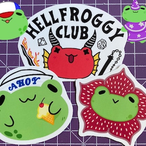 May include: Five frog stickers with different designs. One frog is wearing a sailor hat and holding an ice cream cone. Another frog is wearing a wizard hat and holding a wand. The third frog is wearing a baseball cap. The fourth frog is a red devil with horns. The fifth frog is inside a red flower.