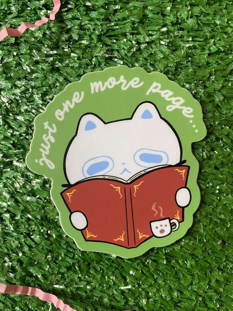 May include: A sticker featuring a white cat reading a red book with the text "just one more page..." on a green background. A small white coffee cup with steam is next to the book. The sticker is on a green surface.