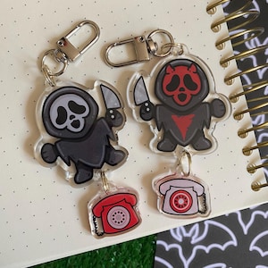 May include: Two acrylic keyrings featuring cartoon figures with knives, one in black and grey, the other in black and red. Each keyring has a red or pink rotary phone charm and a silver clasp. The keyrings are on a notebook.
