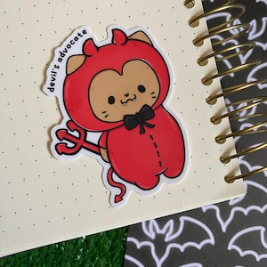 Puede incluir: Una pegatina con un personaje de dibujos animados disfrazado de diablo. El personaje es marrón y lleva un disfraz de diablo rojo, una pajarita negra y un pequeño tridente. El texto "devil's advocate" está en una etiqueta blanca.