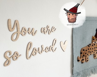 May include: Wooden wall art spelling "You are so loved" in cursive, plus a heart symbol. A blue wall hanging with a leopard design and a brown leather strap is also visible.