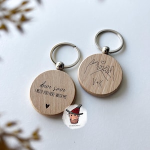 Könnte beinhalten: Zwei Holz-Schlüsselanhänger mit silbernen Ringen. Ein Schlüsselanhänger hat den Text "drive safe I NEED YOU HERE WITH ME" und ein Herz. Der andere hat die Initialen "V+K" und ein Herzsymbol mit verschlungenen Händen.