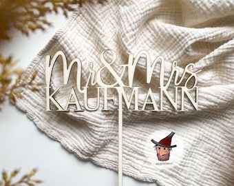以下が含まれることがあります： 「Mr & Mrs KAUFMANN」と書いた、筆記体で書かれた木製ケーキトッパー。トッパーは白い布の背景の上に置かれています。