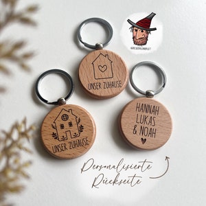 Könnte beinhalten: Drei Holzschlüsselanhänger mit silbernen Ringen. Zwei Schlüsselanhänger haben eine Hausgrafik und die Worte "UNSER ZUHAUSE". Der dritte Schlüsselanhänger trägt die Namen "HANNAH LUKAS & NOAH" und ein Herz. Eine Cartoon-Illustration befindet sich oben rechts.