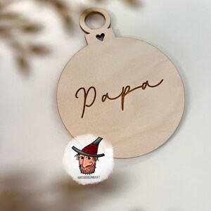 May include: A wooden Christmas ornament shaped like a bauble with the word "Papa" engraved on it. The ornament has a small heart cutout at the top.
