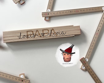 May include: A wooden ruler with the words "Louis & Livia Papa" engraved on it. The ruler is surrounded by other rulers and a cartoon drawing of a person with a beard and a hat.