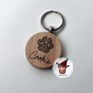 Könnte beinhalten: Ein Holz-Haustieranhänger mit einem silbernen Schlüsselring. Der runde Anhänger hat einen Pfotenabdruck mit Leopardenmuster und den Namen "Cookie" in Schreibschrift. Rechts befindet sich eine kleine Cartoon-Illustration.