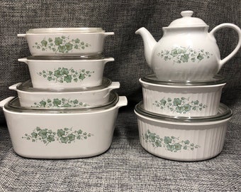 Puede incluir: Un juego de utensilios de cocina de cerámica blanca con un diseño floral verde. El juego incluye una tetera y varios platos para hornear con tapas.