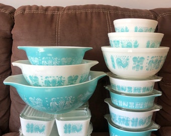 Może przedstawiać: Kolekcja misek i pojemników Pyrex w stylu vintage w różnych rozmiarach. Miski mają turkusowo-białą kolorystykę z dekoracyjnym wzorem przedstawiającym ludzi, koguty i rośliny. Niektóre mają przezroczyste pokrywki.