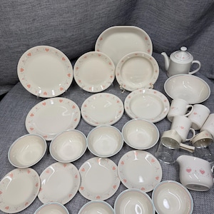 May include: A collection of cream-colored ceramic dinnerware featuring a delicate pattern of small pink hearts and thin green lines. The set includes various plates, bowls, mugs, a teapot, and a creamer, all arranged together on a textured gray surface.