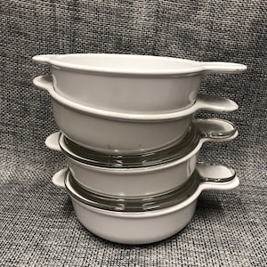 May include: A stack of four white ceramic bowls with glass lids. The bowls have rounded edges and handles on the sides. The bowls are stacked on a gray textured surface.