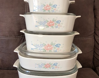 Op de afbeelding: Een set van zes witte ovenschaaltjes met bloemmotieven in roze, blauwe en groene tinten. De schaaltjes zijn op elkaar gestapeld, met het grootste schaaltje onderaan en het kleinste schaaltje bovenaan.