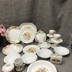 Puede incluir: Un conjunto de vajilla de cerámica blanca con un diseño floral. El juego incluye platos, cuencos, tazas, platillos, una jarra de crema, un azucarero y una fuente. El patrón floral presenta flores amarillas, moradas y rojas con detalles verdes.