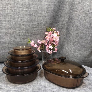 Könnte beinhalten: Ein Satz brauner Glasbackformen mit Deckeln. Die Formen sind übereinander gestapelt und haben ein geripptes Design. Die Formen befinden sich auf einer grauen, strukturierten Oberfläche.