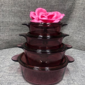 Puede incluir: Un juego de cuatro fuentes para hornear de vidrio morado apiladas una encima de la otra. Las fuentes son redondas y tienen una tapa de vidrio transparente. Una flor rosa está encima de las fuentes.