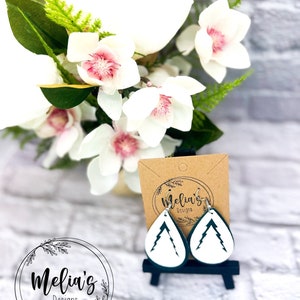May include: A pair of teal green teardrop-shaped earrings with a white silhouette of a mountain inside. The earrings are displayed on a small easel with a brown card that says "Melia's Designs".