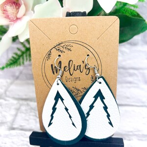 May include: A pair of white leather earrings with a green outline. The earrings are shaped like teardrops and have a white silhouette of a pine tree inside. The earrings are hanging from silver hooks. The earrings are displayed on a brown card with the text "Melia's Designs".