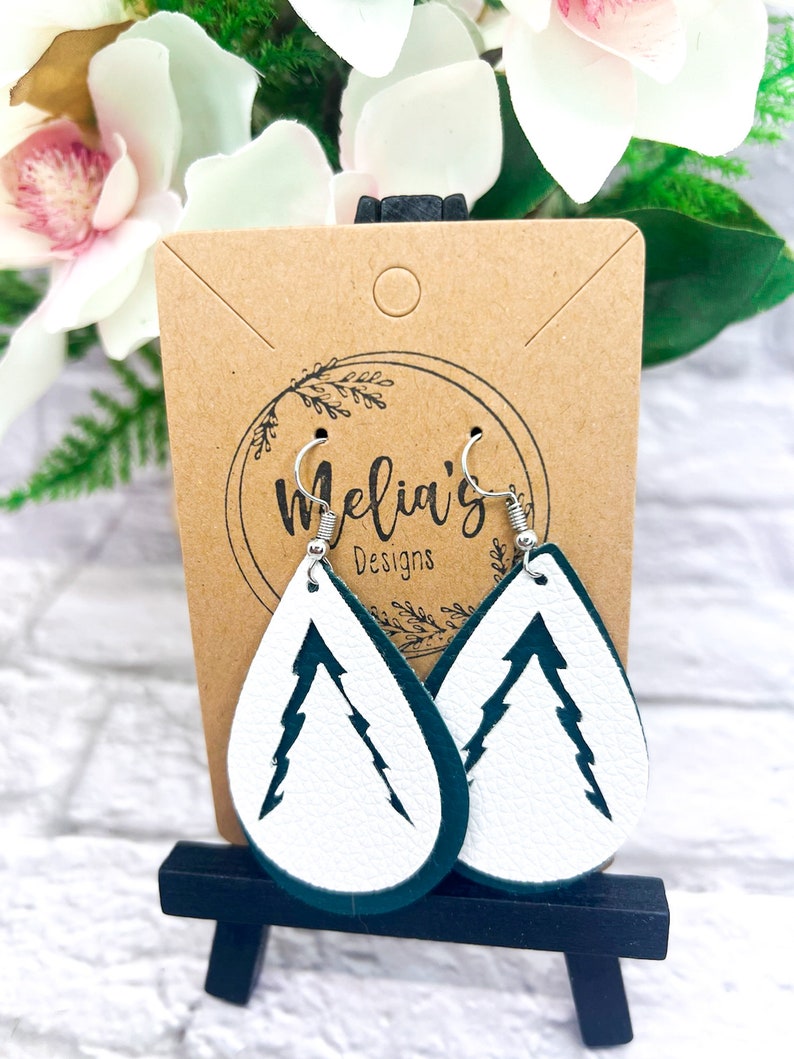 May include: A pair of white leather earrings with a green outline. The earrings are shaped like teardrops and have a white silhouette of a pine tree inside. The earrings are hanging from silver hooks. The earrings are displayed on a brown card with the text "Melia's Designs".