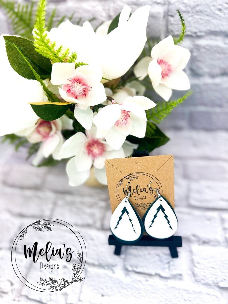 May include: A pair of teal green teardrop-shaped earrings with a white silhouette of a mountain inside. The earrings are displayed on a small easel with a brown card that says "Melia's Designs".