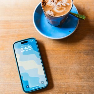 May include: A smartphone with a blue case displaying the time 3:55 and a blue and white wavy pattern. A cup of coffee with latte art sits on a blue saucer with a green spoon on a wooden table.