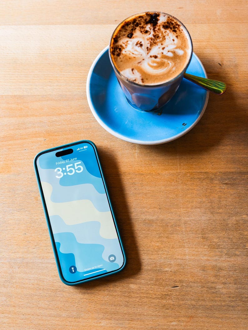 May include: A smartphone with a blue case displaying the time 3:55 and a blue and white wavy pattern. A cup of coffee with latte art sits on a blue saucer with a green spoon on a wooden table.