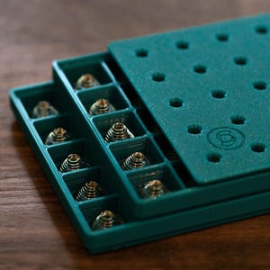 May include: Teal blue plastic storage case with multiple compartments for small items. The case has a textured surface and a logo on the top.