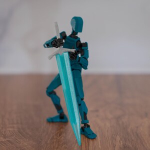 Puede incluir: Una figura de acci&oacute;n articulada de color azul turquesa sosteniendo una espada azul turquesa con una empu&ntilde;adura plateada. La figura est&aacute; de pie sobre una superficie de madera.
