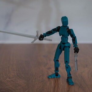 Puede incluir: Una figura de acci&oacute;n articulada de color azul turquesa que sostiene una espada plateada y una pistola plateada. La figura est&aacute; de pie sobre una superficie de madera.
