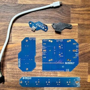 Pode incluir: Um conjunto de placas de circuito azul com texto branco e conectores pretos. As placas são etiquetadas como "Frets On V2" e "Stronger V2". Há também uma peça de plástico preto e um cabo branco com um conector USB.