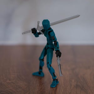 Puede incluir: Una figura de acci&oacute;n azul turquesa sosteniendo una espada y una daga plateadas. La figura est&aacute; de pie sobre una superficie de madera.