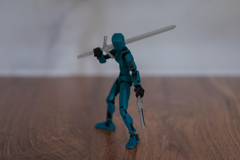 Puede incluir: Una figura de acci&oacute;n azul turquesa sosteniendo una espada y una daga plateadas. La figura est&aacute; de pie sobre una superficie de madera.
