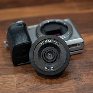 May include: A black 30mm F/11 camera lens with a 49mm diameter, resting in front of a silver digital camera. The lens is centered, with the camera body and black leather grip visible in the background. The setup is on a wooden surface.