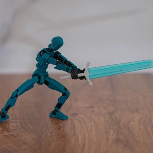Puede incluir: Una figura de acci&oacute;n articulada de color azul verdoso sosteniendo una espada azul transl&uacute;cida con una empu&ntilde;adura plateada. La figura est&aacute; en posici&oacute;n de combate con una pierna hacia adelante y la otra hacia atr&aacute;s. La figura est&aacute; de pie sobre una superficie de madera.