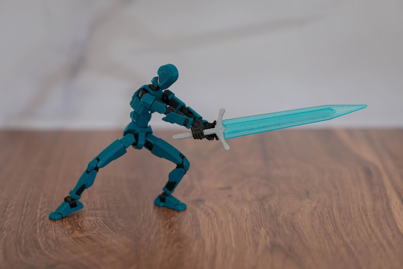 Puede incluir: Una figura de acci&oacute;n articulada de color azul verdoso sosteniendo una espada azul transl&uacute;cida con una empu&ntilde;adura plateada. La figura est&aacute; en posici&oacute;n de combate con una pierna hacia adelante y la otra hacia atr&aacute;s. La figura est&aacute; de pie sobre una superficie de madera.