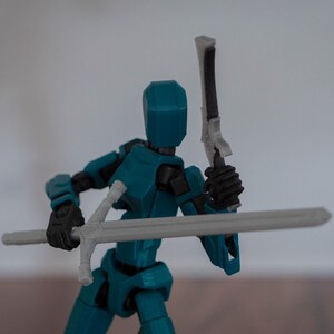 Puede incluir: Una figura de acci&oacute;n articulada de color azul turquesa que sostiene una espada plateada y una espada blanca y negra. La figura tiene una cabeza azul y manos negras.