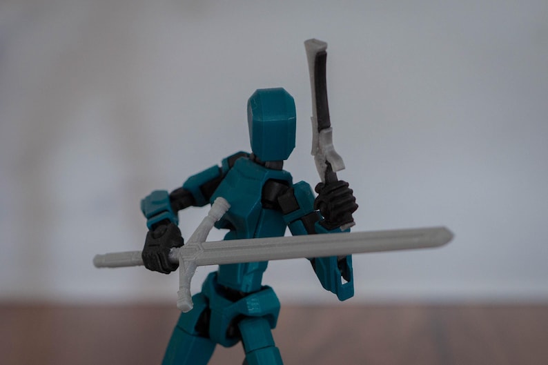 Puede incluir: Una figura de acci&oacute;n articulada de color azul turquesa que sostiene una espada plateada y una espada blanca y negra. La figura tiene una cabeza azul y manos negras.