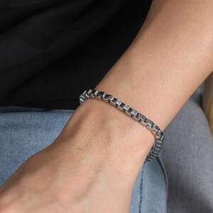 May include: A silver box chain bracelet made of metal. The bracelet is worn on a wrist. The background shows a person wearing a black top and blue jeans.
