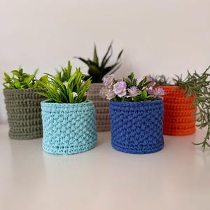 May include: A selection of handmade crochet plant pots in shades of blue, orange, and green. Each pot contains plants or flowers. The pots are displayed on a white surface. Measurements are in centimetres.