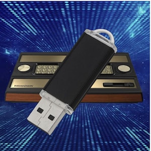 May include: A black USB flash drive with a clear loop is angled in front of a vintage Intellivision game console. The console has a wood grain base, gold accents, and a keypad. The background is a blue digital pattern.