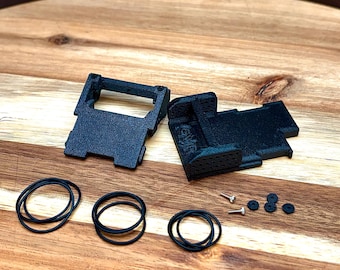 May include: Two black 3D-printed plastic mounting brackets with textured surfaces, accompanied by three black rubber O-rings, two small metal screws, and four small black circular washers, all arranged as individual components.