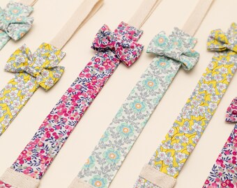 May include: A collection of fabric hair accessories featuring long, flat, floral-patterned ribbons in yellow, pink, and light blue, each topped with a matching small fabric bow and attached to a neutral-colored elastic band.